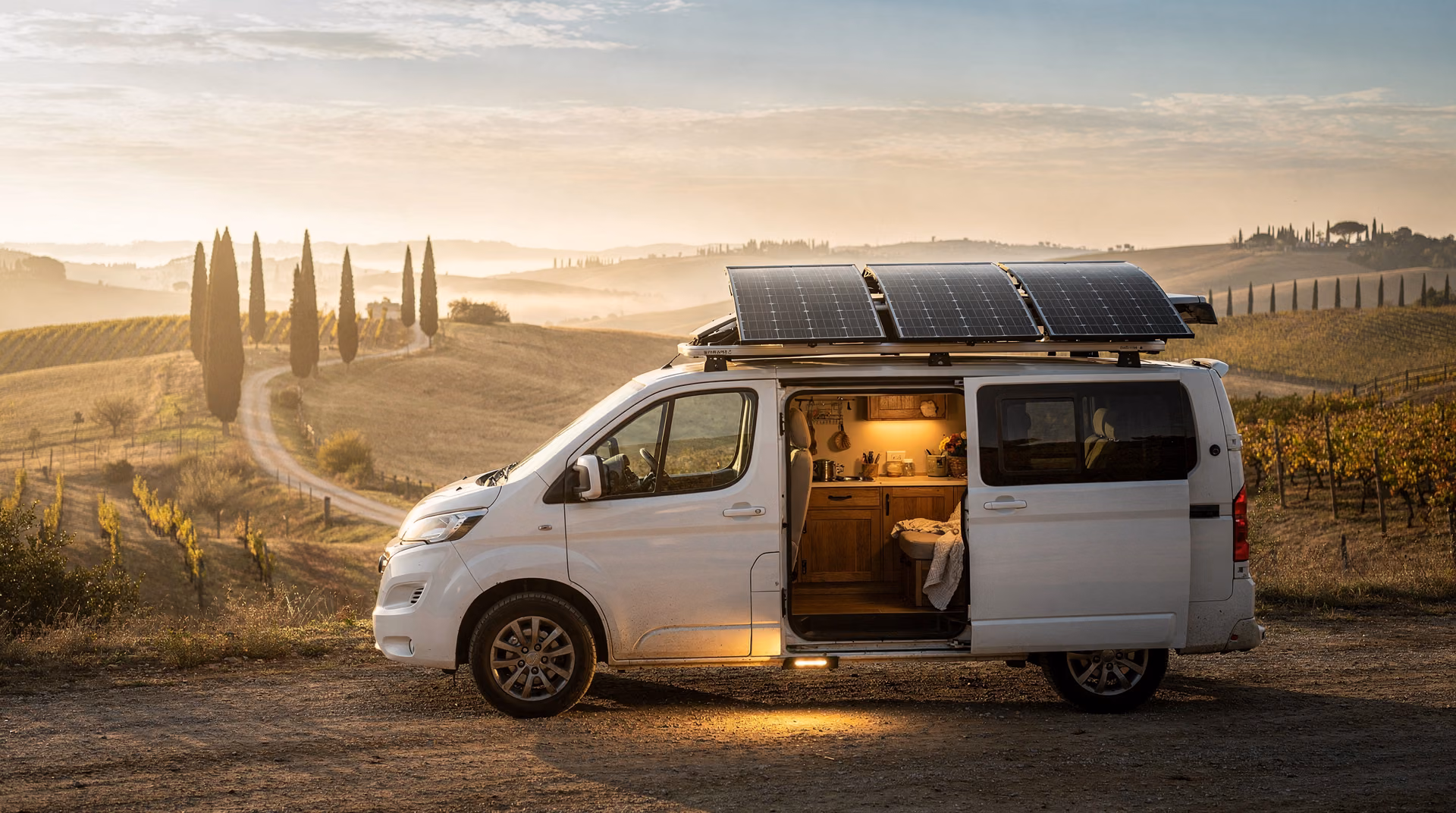 Camper bianco parcheggiato in paesaggio toscano con pannelli solari sul tetto e porta laterale aperta